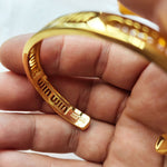 Gold openwork logo bangle close-up, engraved logo text along the open cuff edge