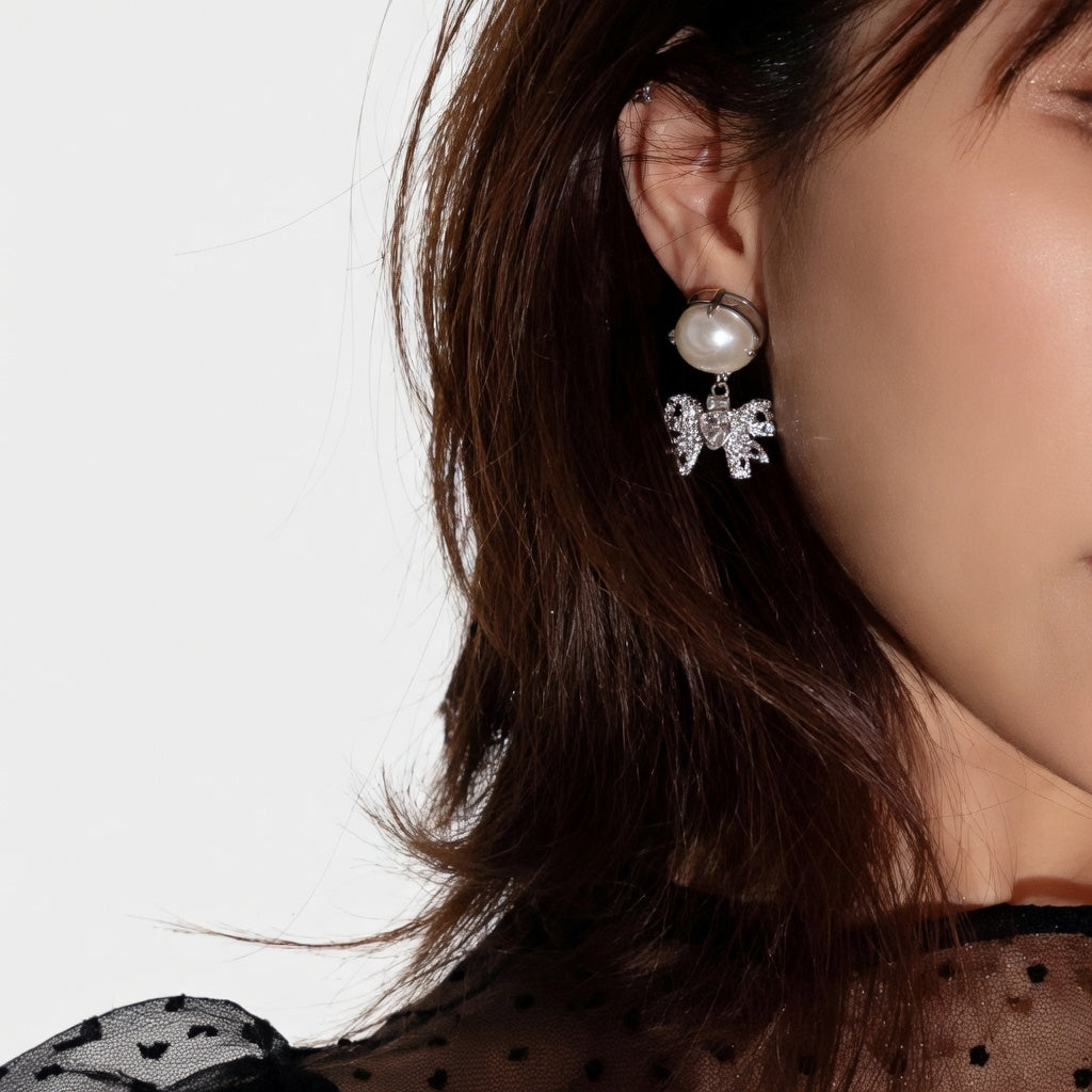 Crystal bow dangle earrings with white pearl on silver-tone studs, close-up.
